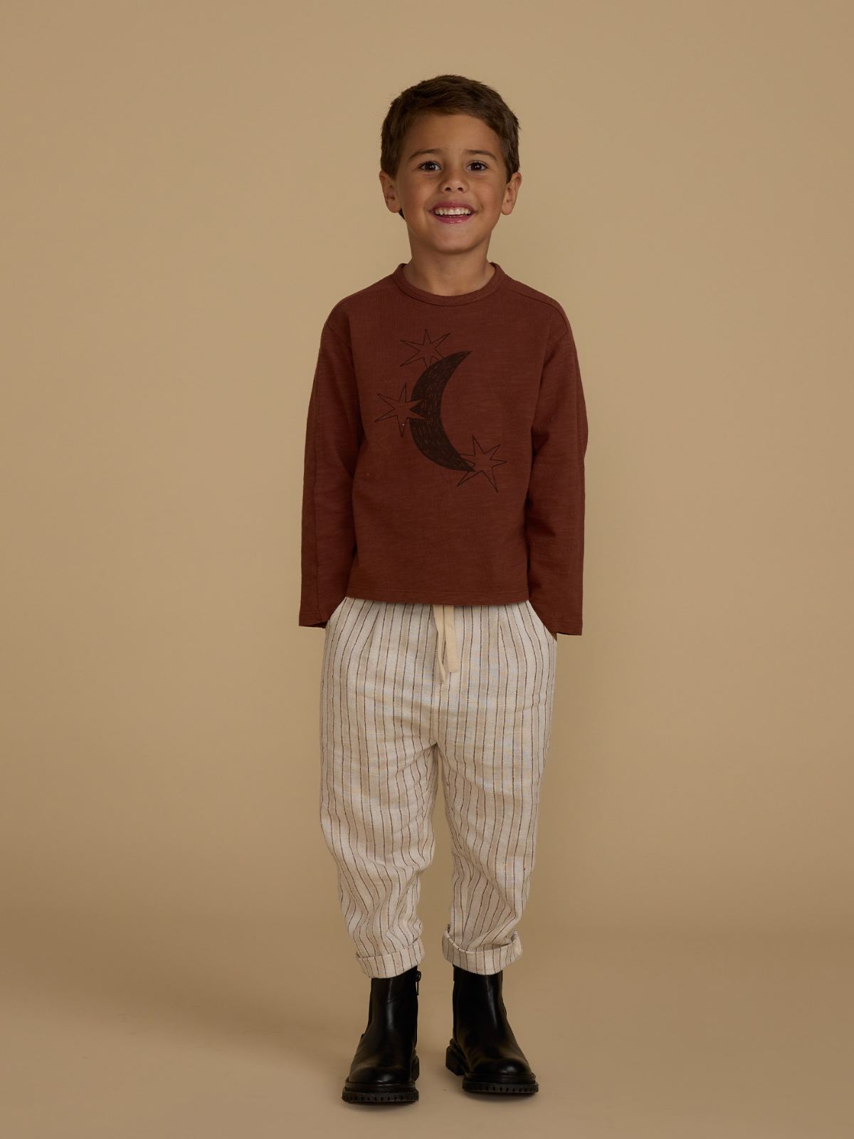 Smiling boy wearing a brown moon-graphic sweatshirt, striped pants, and black boots, against a beige background.