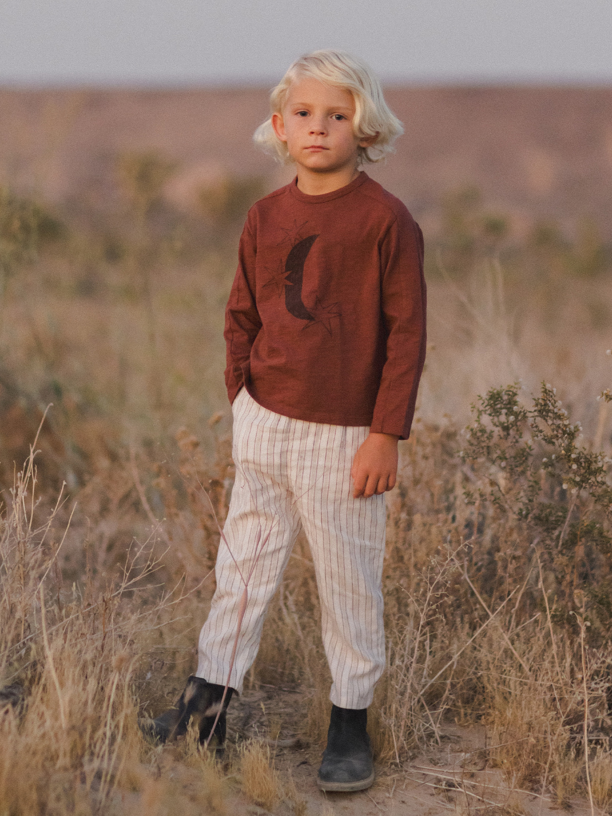 Child with curly blonde hair wearing a maroon shirt and striped pants, standing in a field at sunset.
