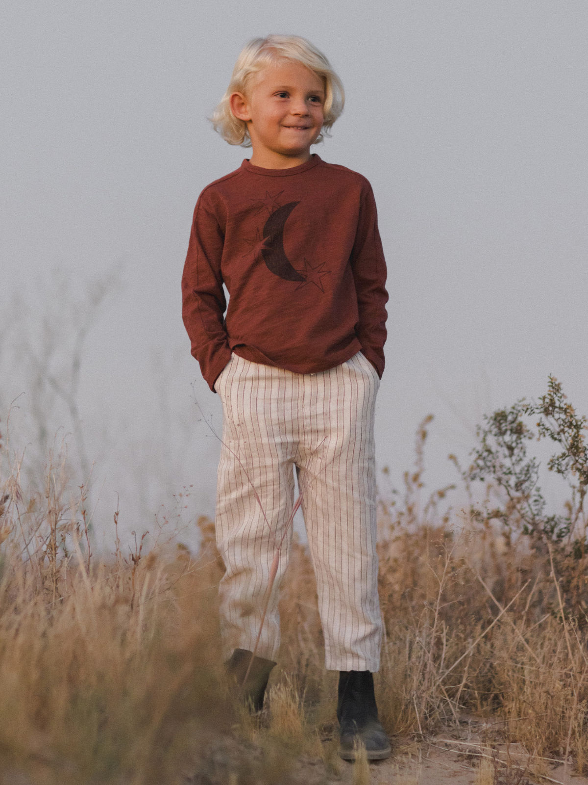 Child with short blonde hair in a brown shirt and striped pants stands outdoors, smiling in a grassy field at dusk.