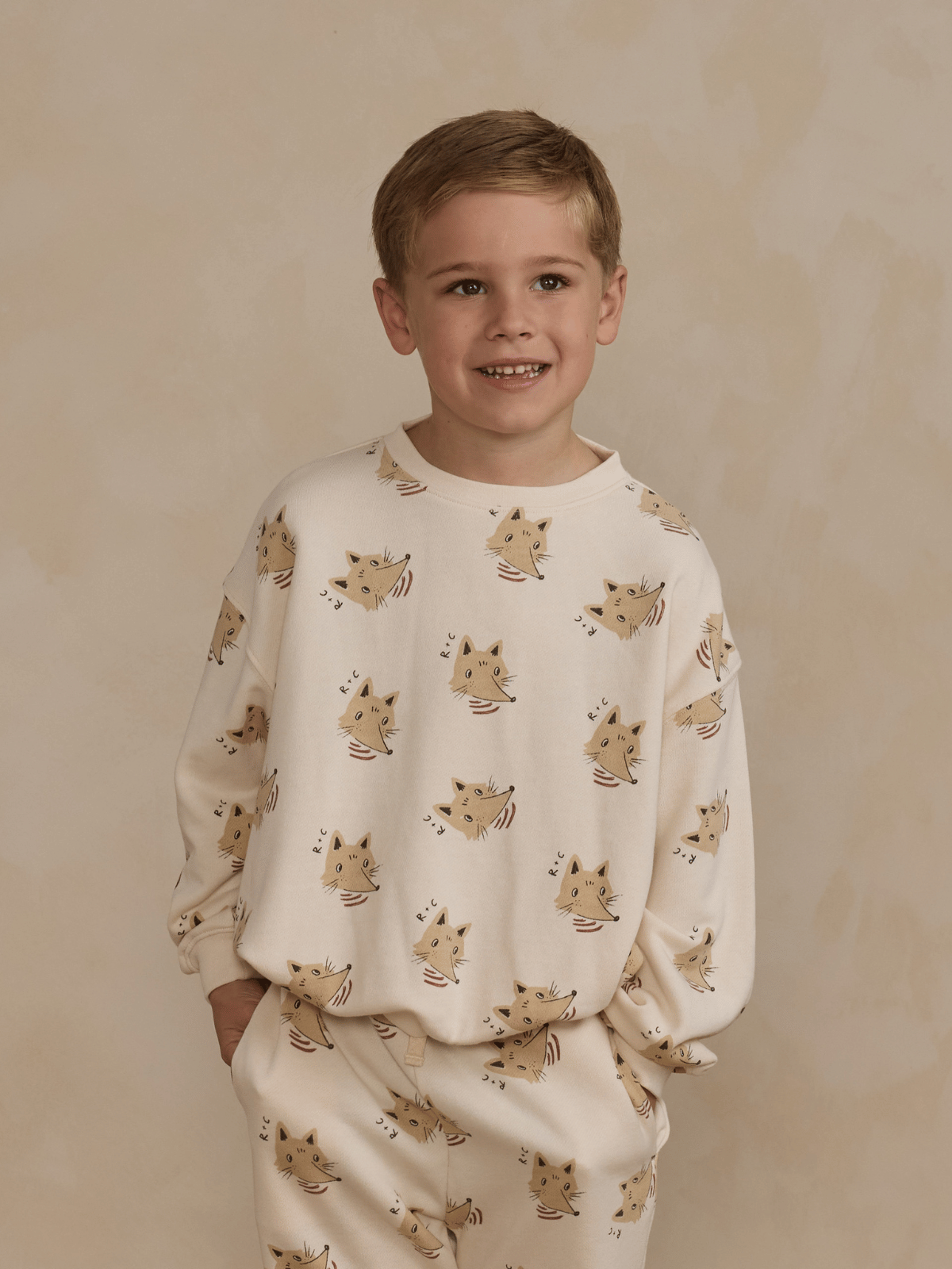 Smiling boy wearing a playful cat-patterned sweatshirt and pants against a neutral background.