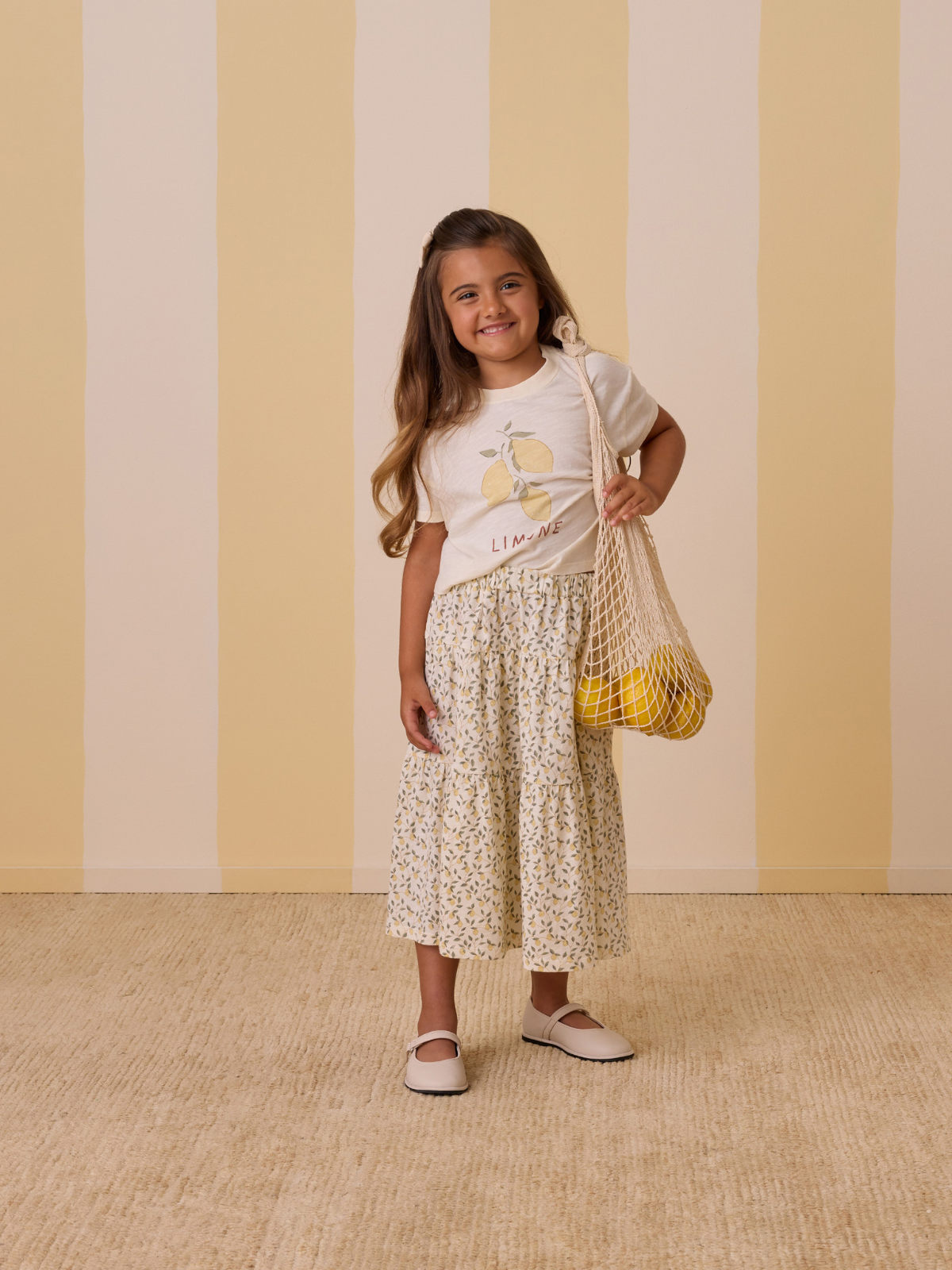 Girl smiling in a lemon-themed shirt and floral skirt, holding a net bag with lemons, against a striped background.