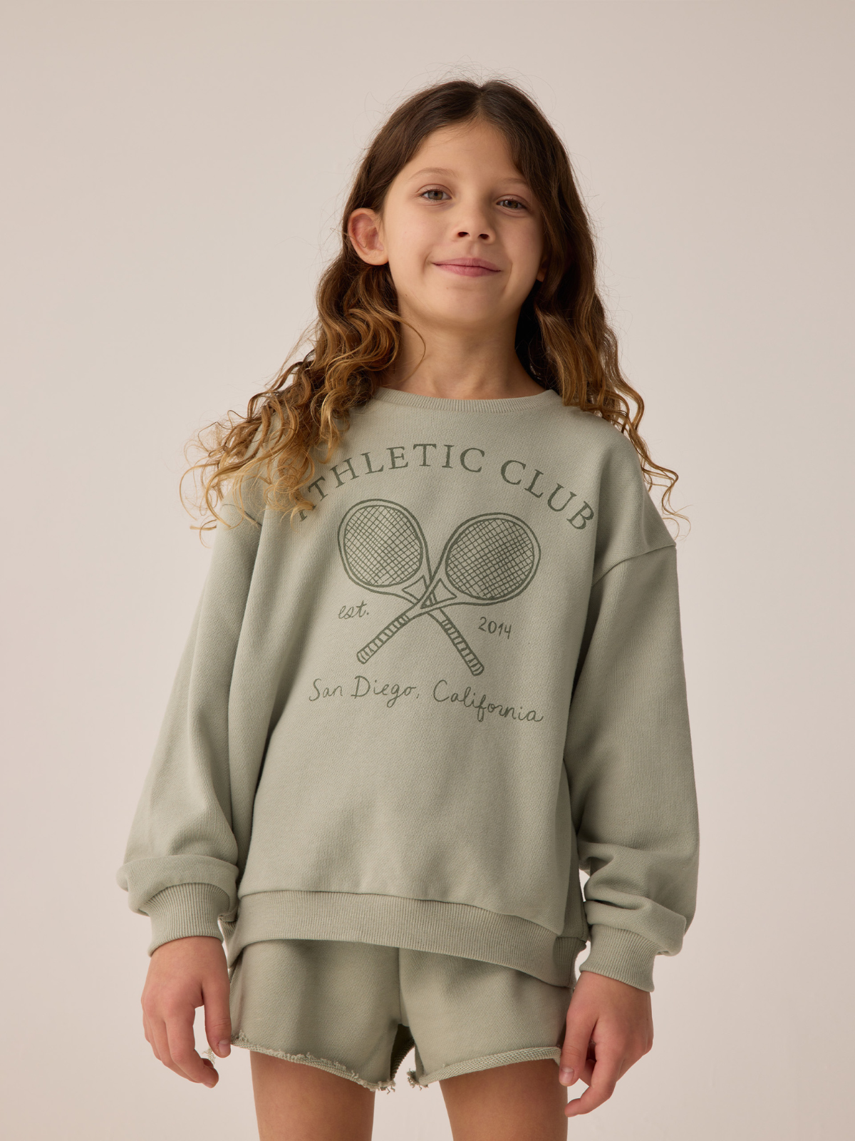 Child wearing a green sweatshirt with tennis racket graphics and "Athletic Club" text, smiling against a neutral background.