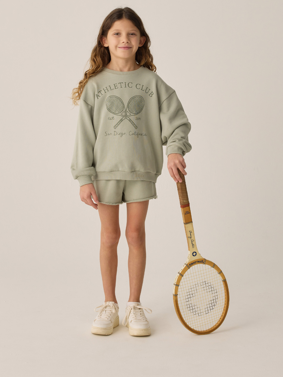 Girl in a green athletic outfit holding a tennis racket, smiling against a neutral background.