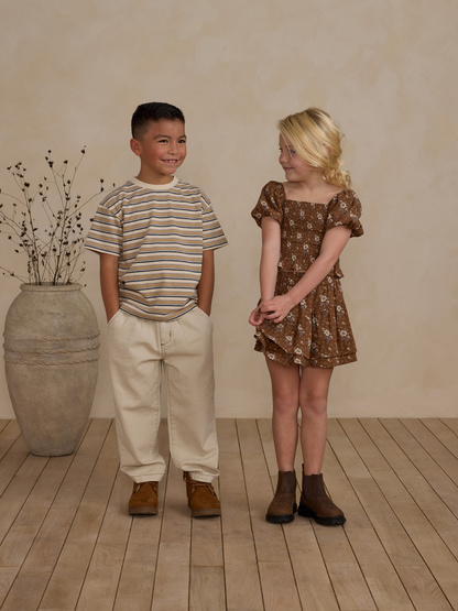 Two children smiling: a boy in a striped shirt and a girl in a floral dress, standing on wooden flooring.
