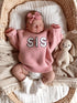 Baby girl in a pink "SIS" sweater sleeps in a basket, with a soft bunny toy beside her. Cozy and peaceful setting.