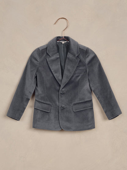 Gray velvet blazer hung on a copper hanger against a textured beige background.
