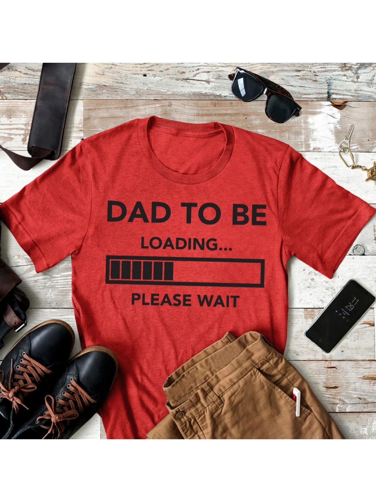 Red t-shirt with "Dad to be loading... Please wait" text, surrounded by sunglasses, shoes, and pants on a wooden surface.