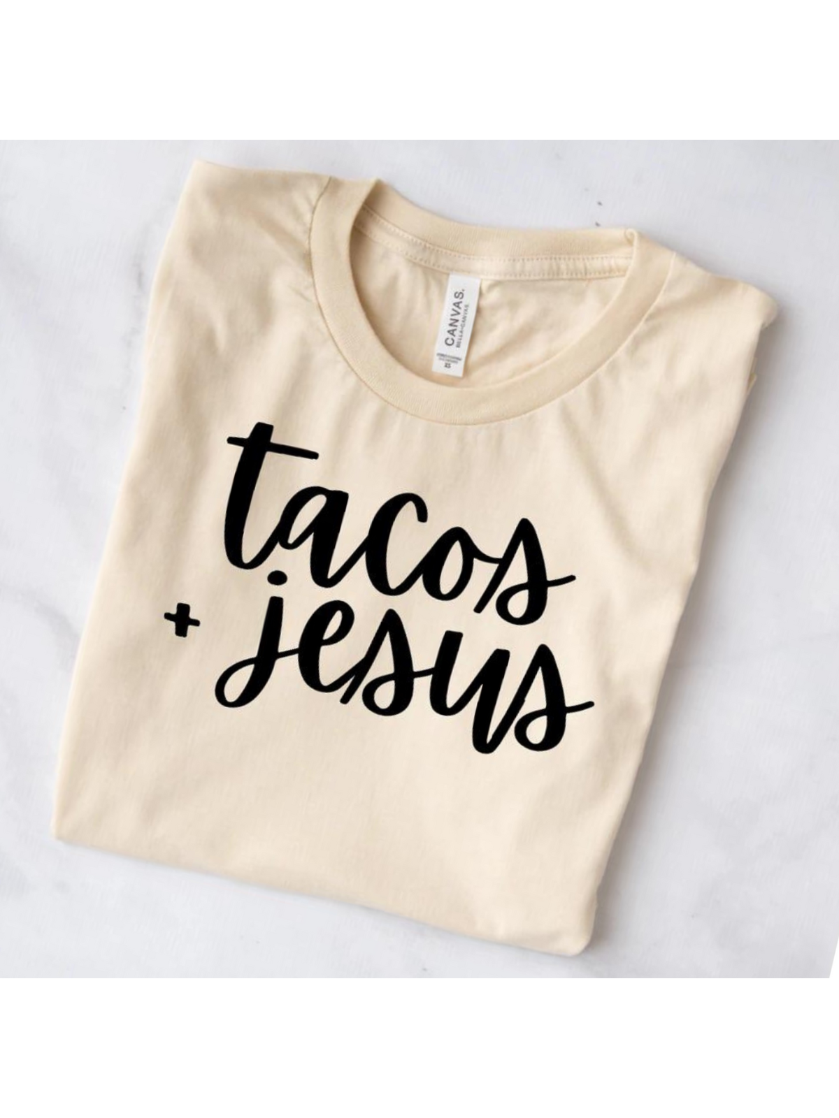 Beige t-shirt with black text saying "tacos + Jesus" displayed on a light surface.