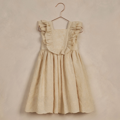 Light beige children's dress with ruffled straps hanging on a copper hanger against a textured background.