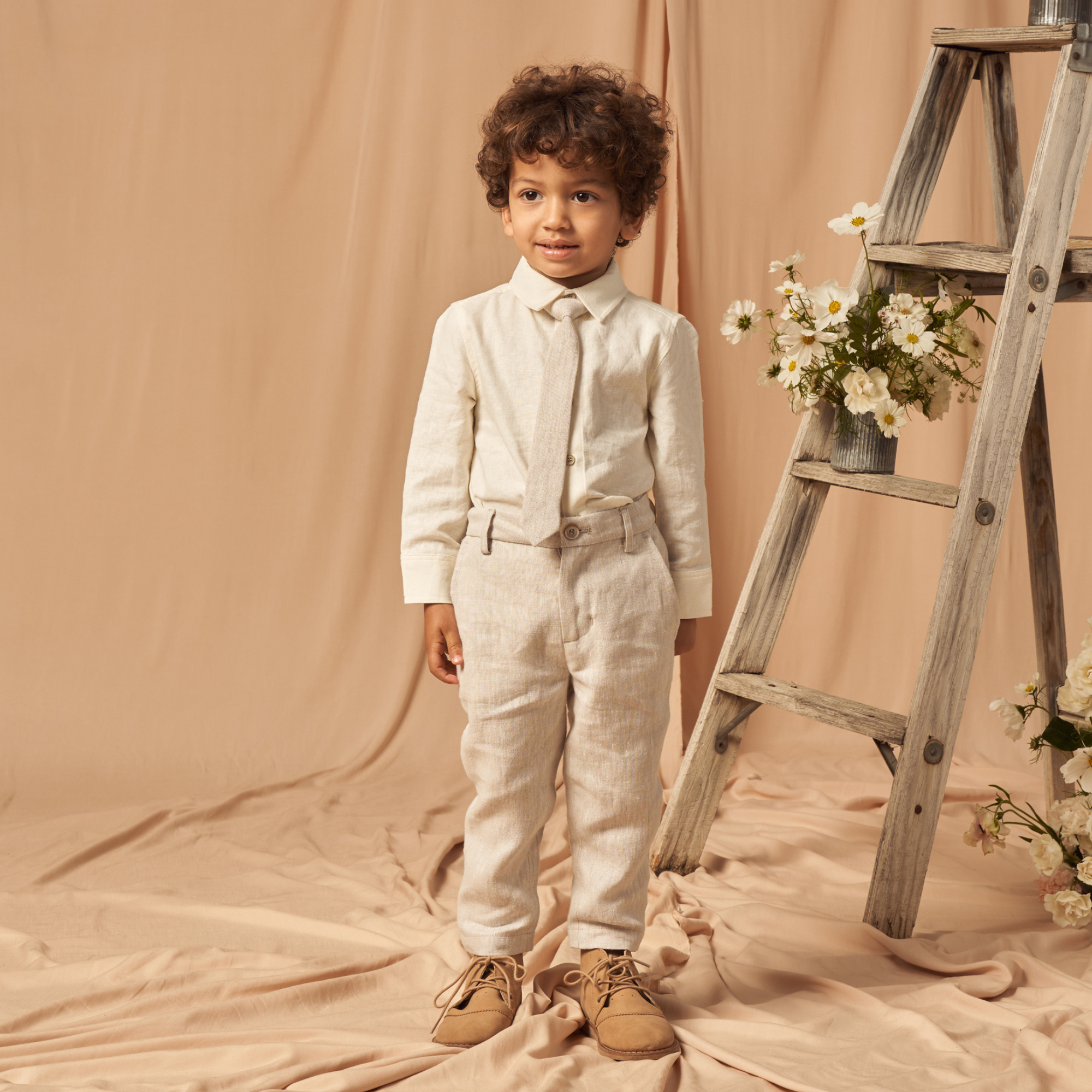 Young boy in a cream suit with a tie, standing beside a decorative ladder and flowers against a beige backdrop.