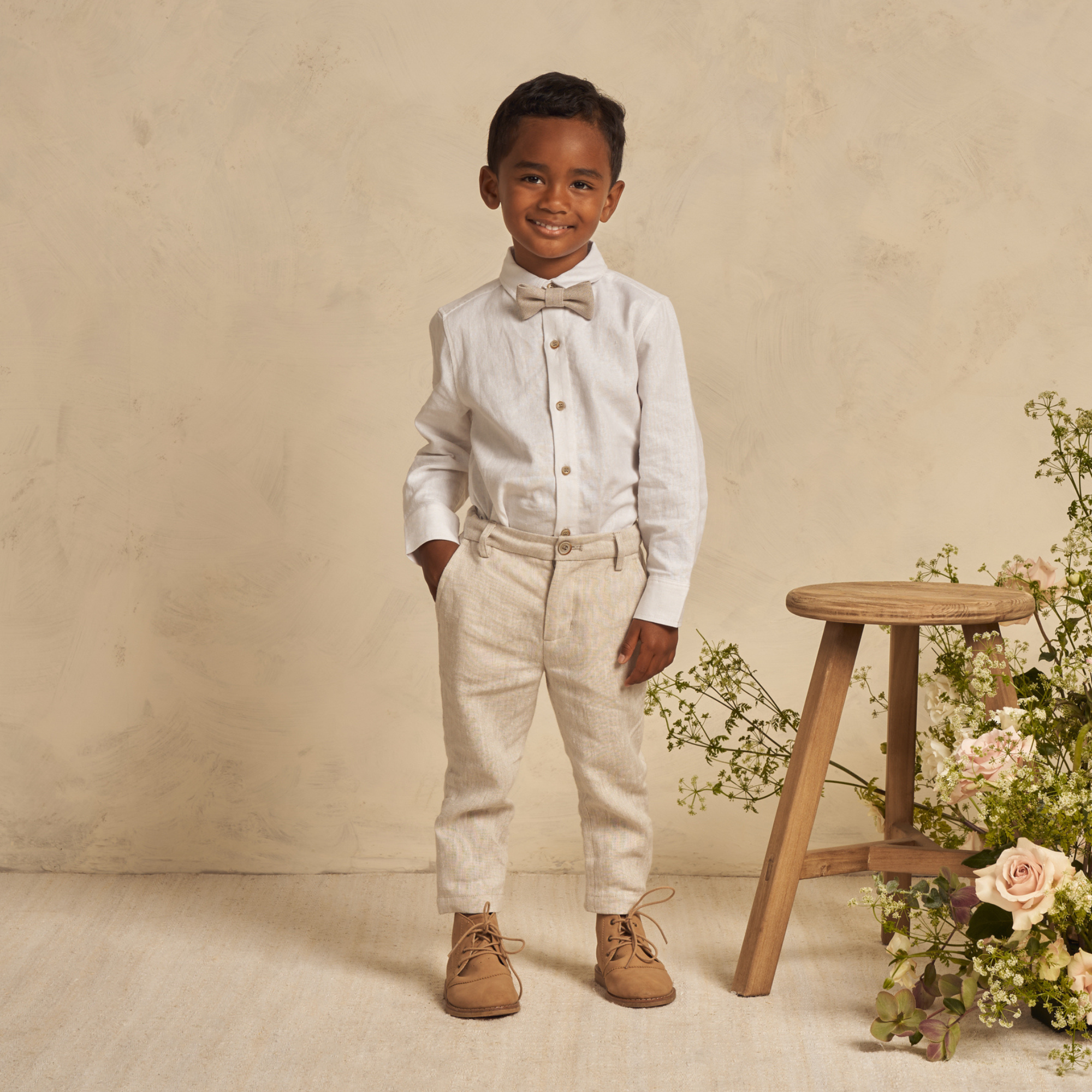 Smiling boy in a white shirt and bow tie, standing beside a wooden stool with flowers in a neutral background.