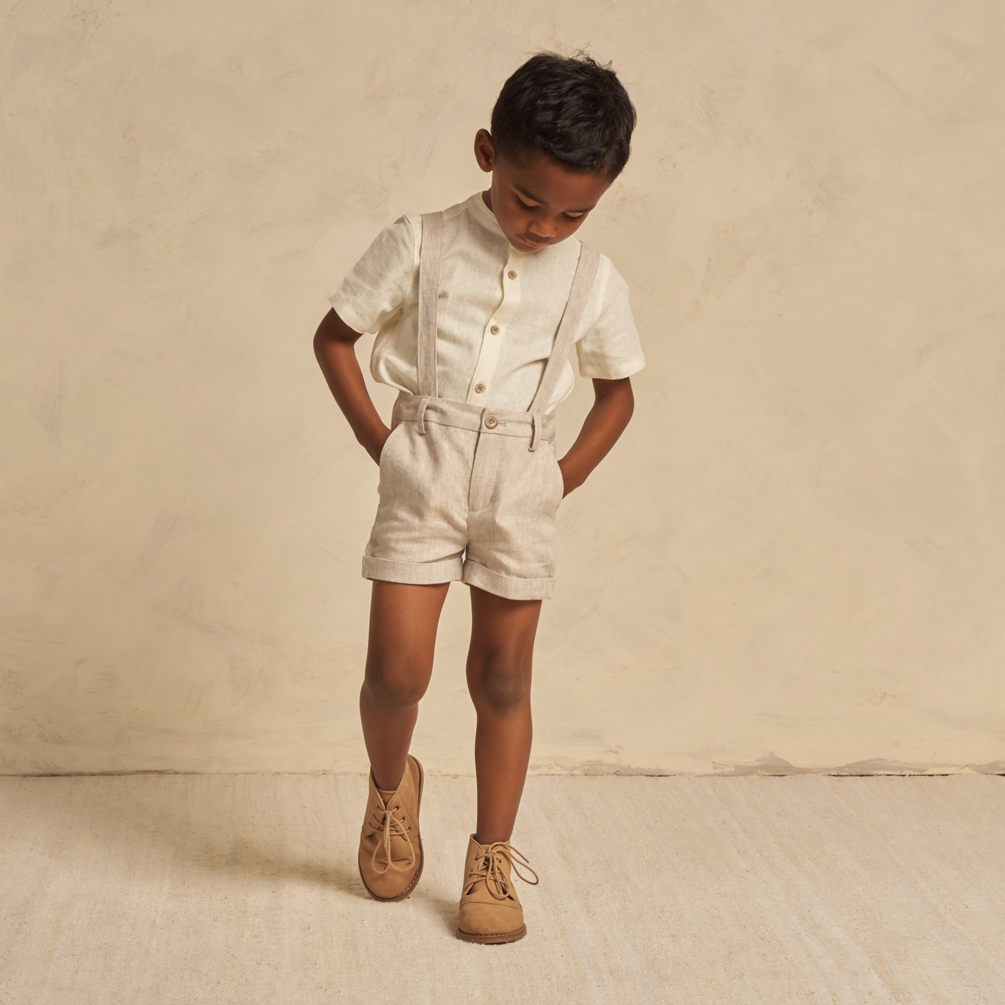 Child in light-colored outfit with suspenders, shorts, and brown shoes, looking down against a textured beige background.