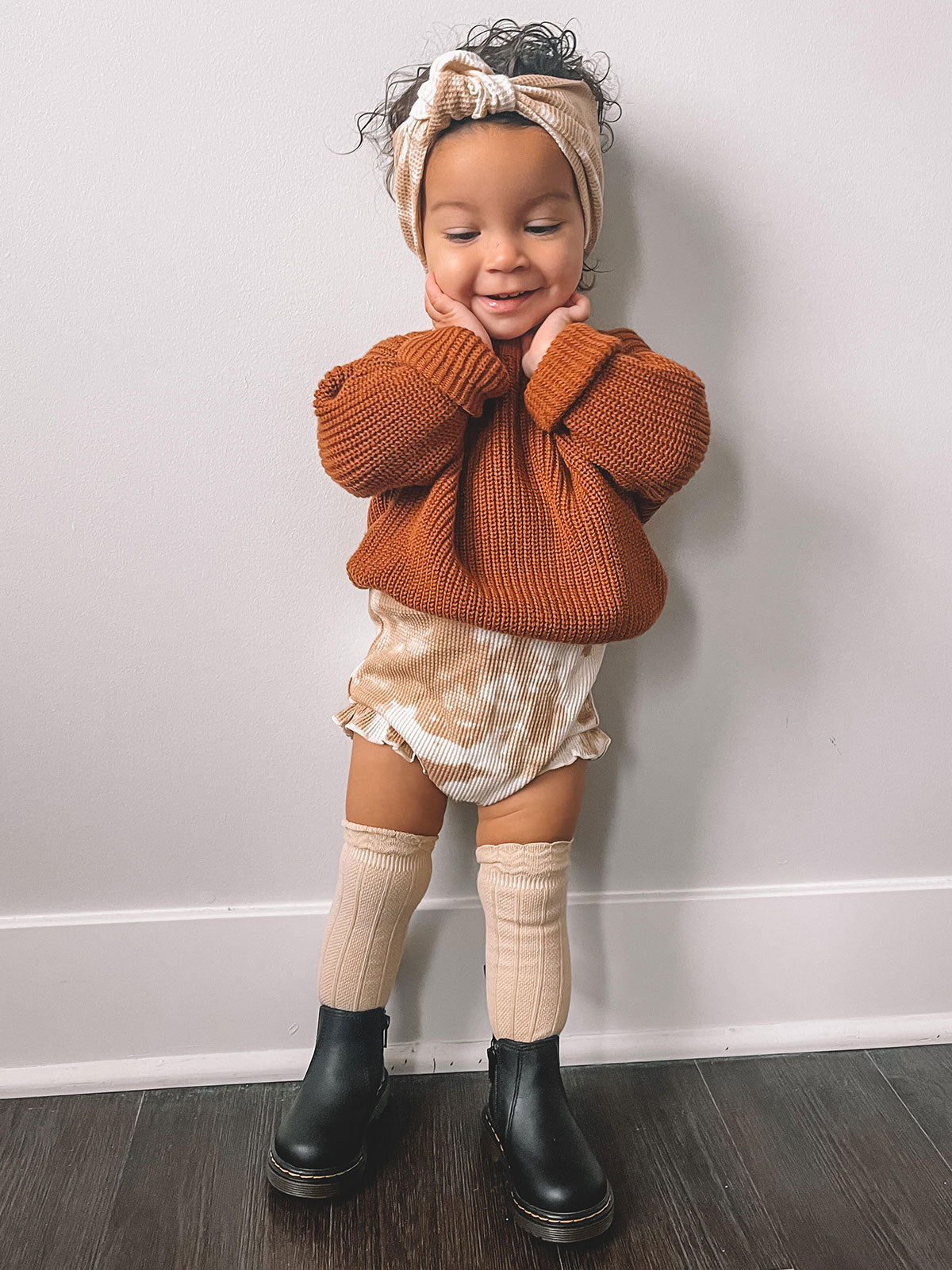 Toddler in a cozy orange sweater and headband, smiling with hands on cheeks, wearing shorts and knee-high socks.