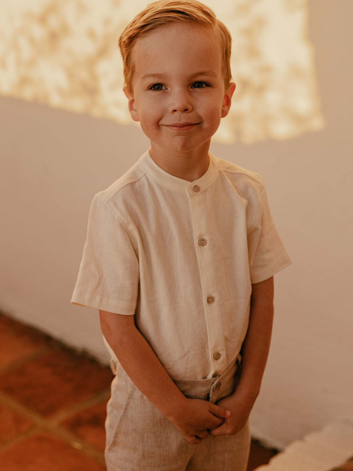 Smiling boy in a beige linen outfit stands with hands clasped, against a softly lit backdrop.