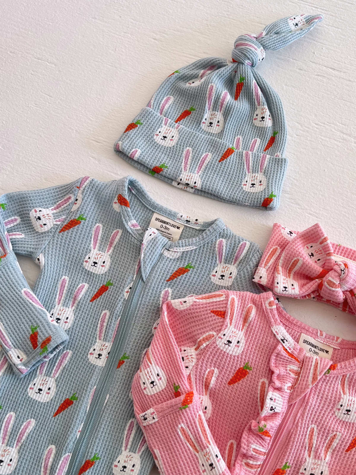 Clothing set featuring bunny and carrot patterns: a blue onesie, pink outfit, hat, and headband.