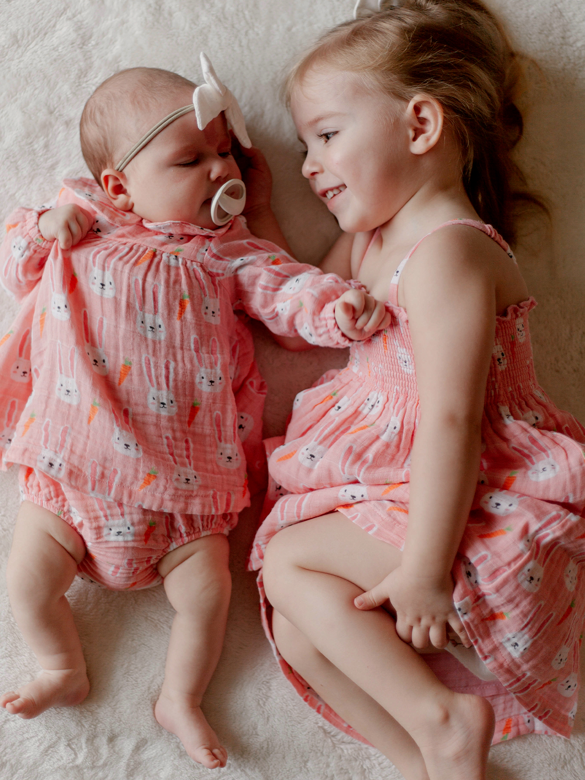 Two young sisters in matching pink bunny print outfits share a joyful moment on a soft blanket.