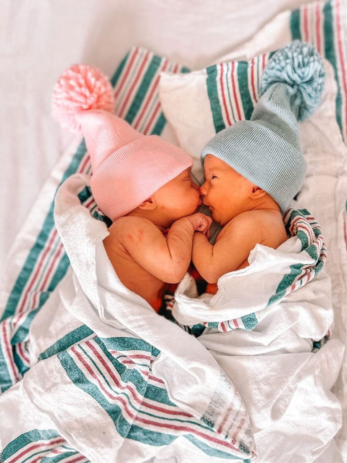 Cute twin babies, one in a pink hat and the other in a blue hat, cuddling under a striped blanket.