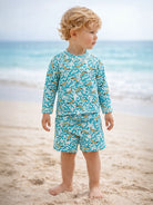 Child in a banana-patterned swimsuit stands on the beach, with ocean waves in the background.