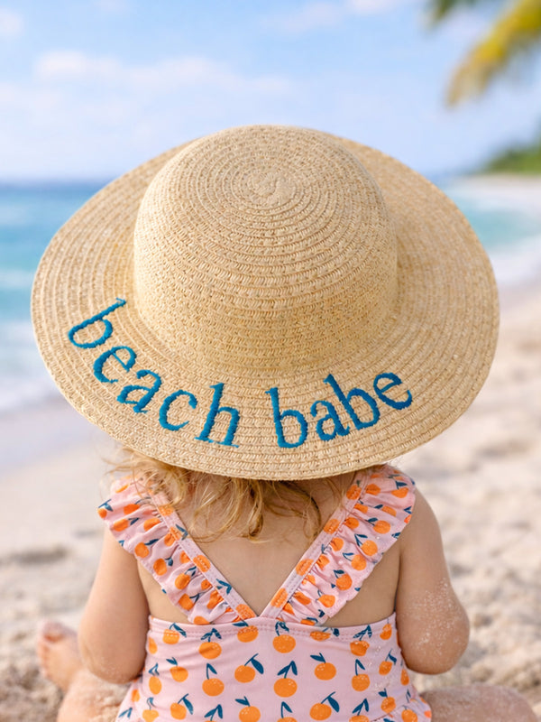 Child in a straw hat reading "beach babe," sitting on a sandy beach with ocean waves in the background.