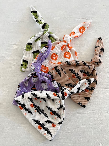 Four colorful baby hats with Halloween-themed patterns featuring pumpkins, bats, and ghosts, tied at the top.