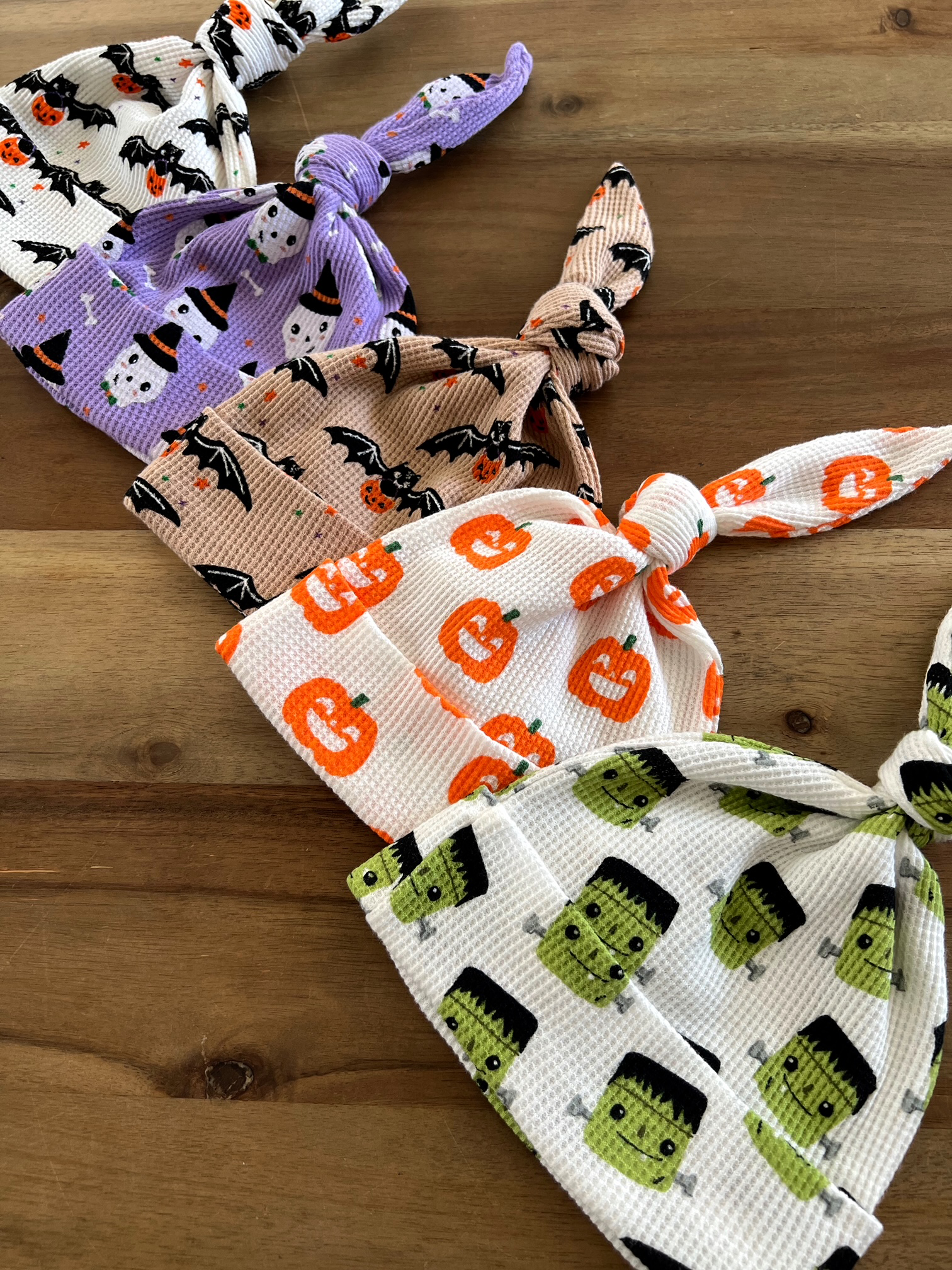 Four Halloween-themed baby hats featuring ghosts, bats, pumpkins, and Frankenstein designs on a wooden surface.