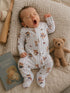 Yawning baby wearing a striped onesie with teddy bear print, surrounded by a book and a plush teddy bear.