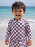 Smiling toddler in a checkered swimsuit on the beach, with ocean waves in the background.