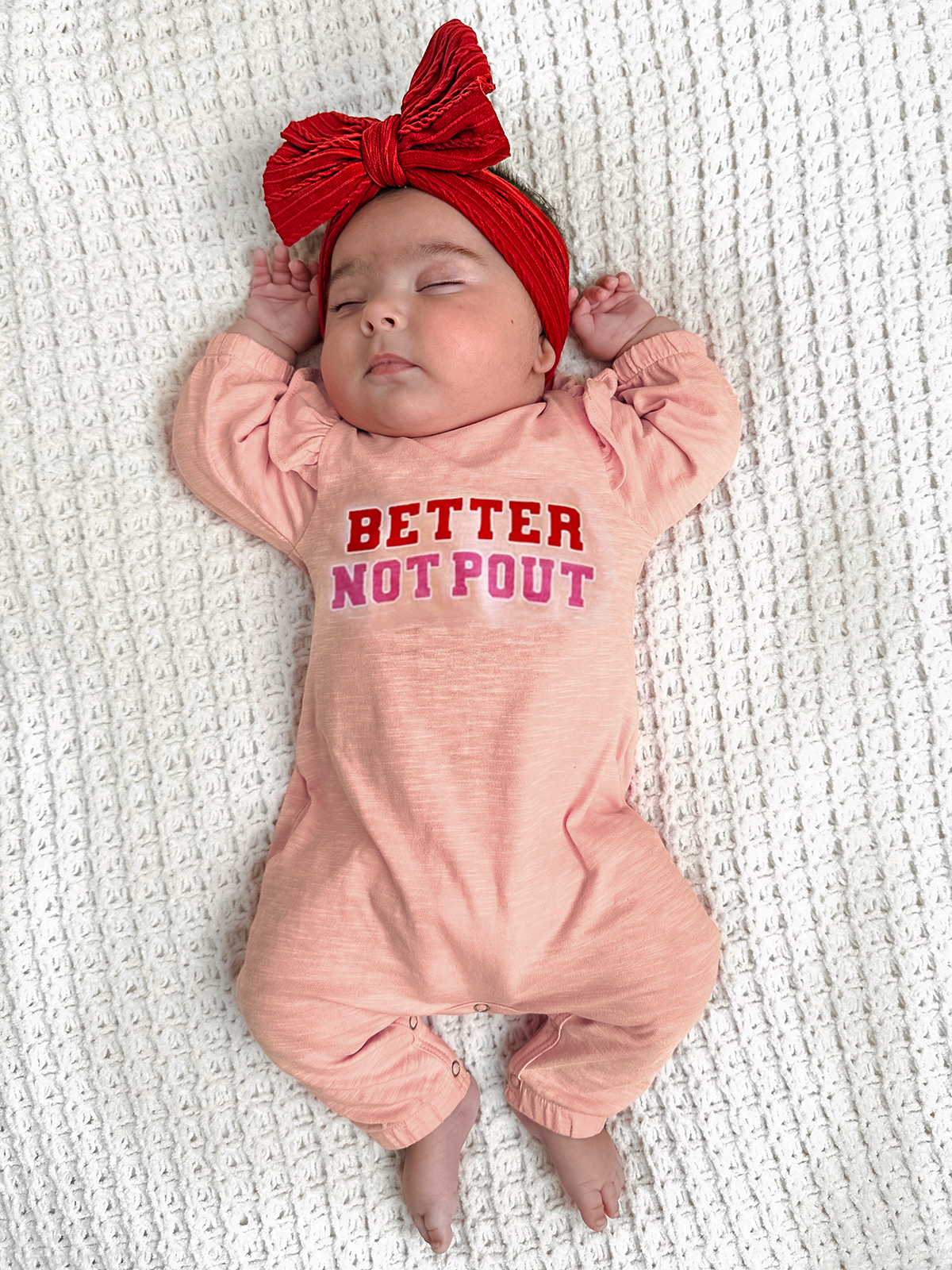 Baby wearing a pink onesie with "Better Not Pout" text, relaxing on a white textured blanket, with a red headband.