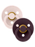 Two pacifiers in beige and dark purple with gold accents, featuring multiple holes for airflow and brand logo.