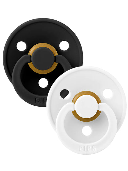 Two pacifiers from BIBS: one black with gold accents, one white with gold accents, placed side by side on a white background.