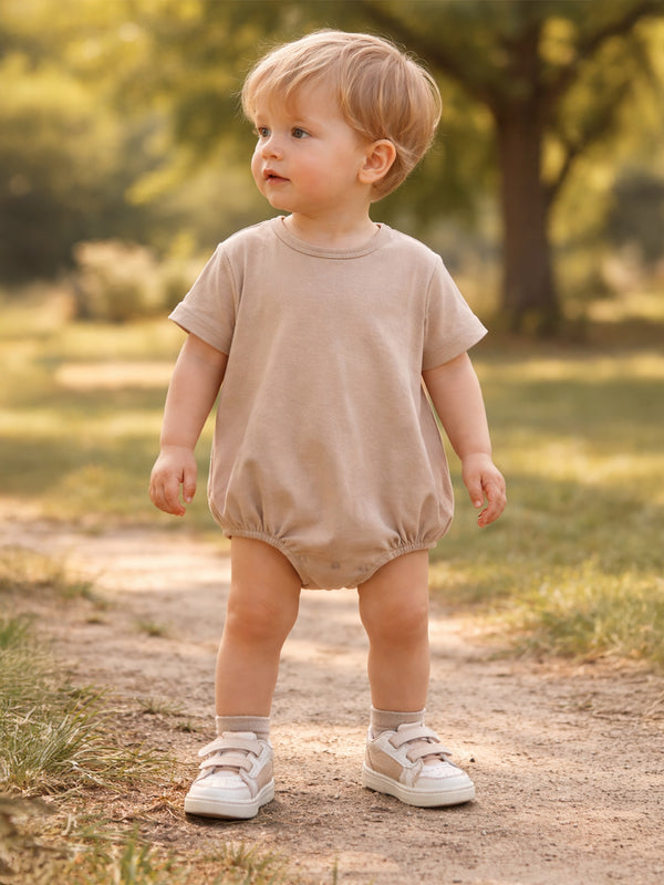 Toddler in a beige romper and sneakers stands on a path in a sunny, green park.