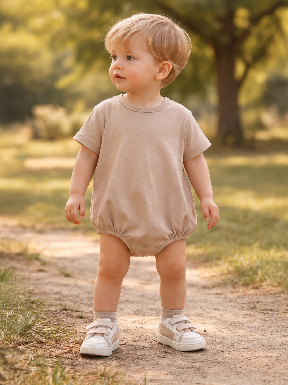Toddler in a beige romper and sneakers stands on a path in a sunny, green park.