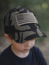Child wearing a camo baseball cap with an American flag patch, looking down outdoors.