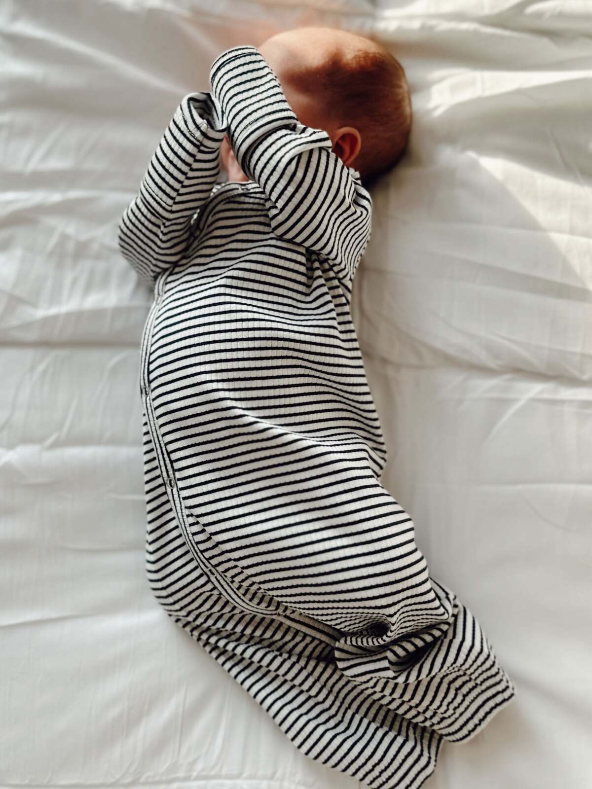 Baby in a striped outfit sleeping on a white bed, arms raised over their face.