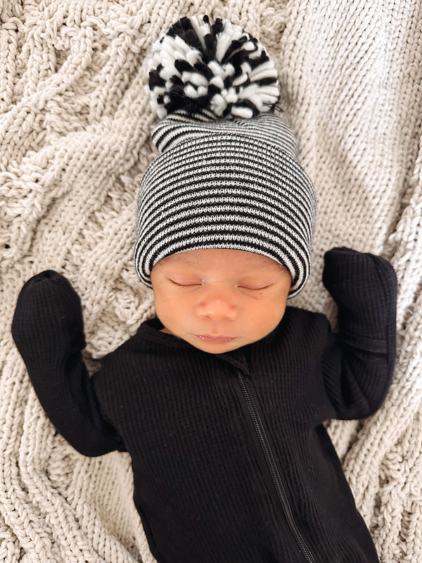 Newborn in striped black and white beanie with pompom, resting on a textured cream blanket.