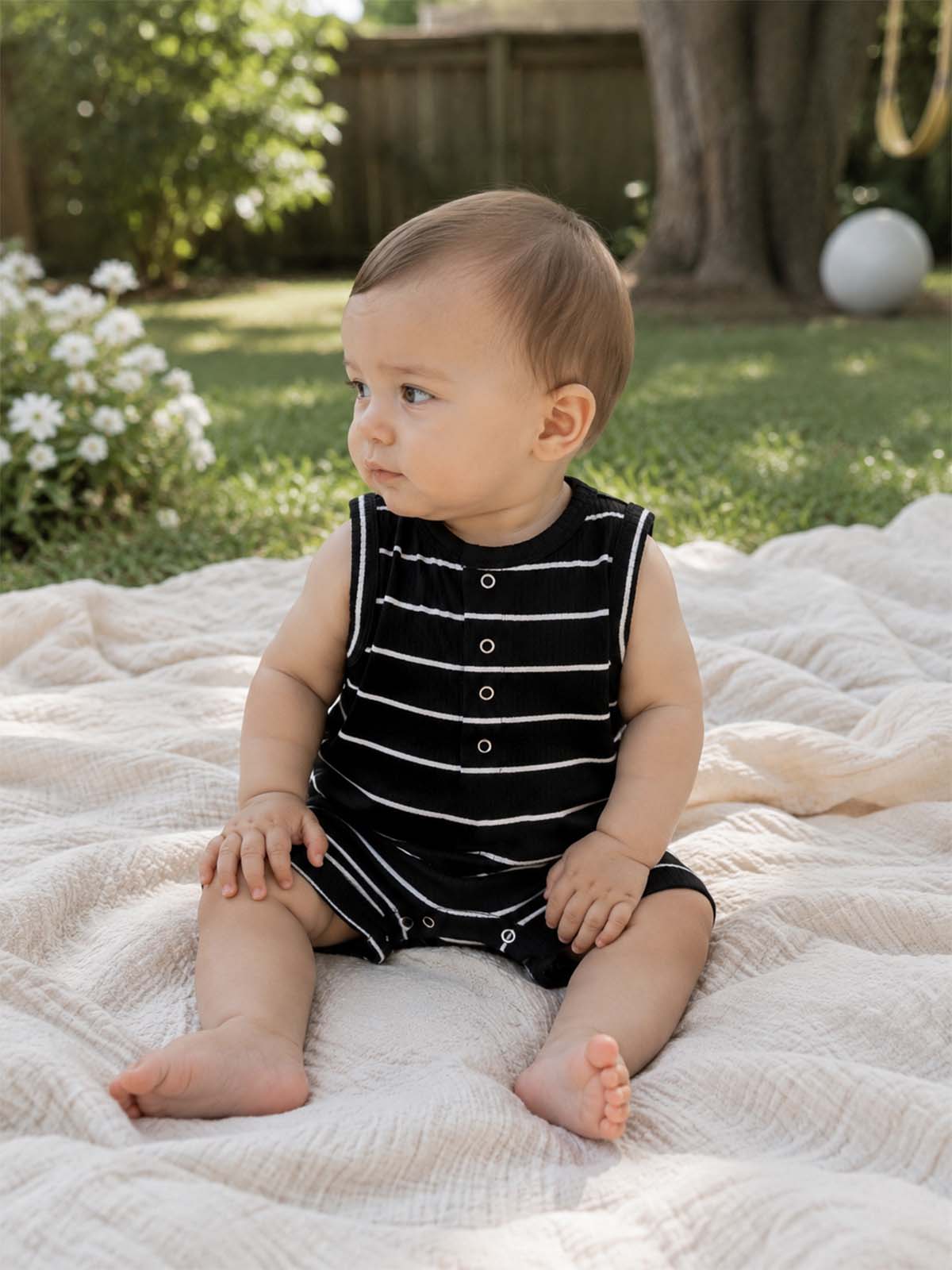 Baby in black and white striped romper sits on a soft blanket outdoors, surrounded by lush green grass and flowers.