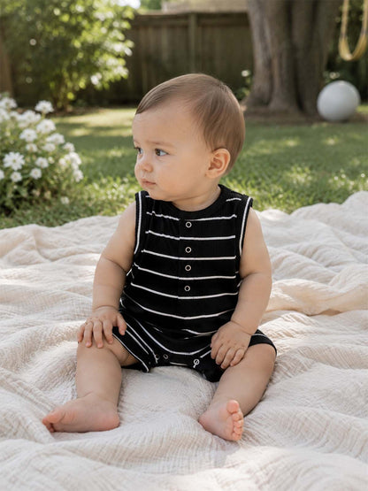Baby in black and white striped romper sits on a soft blanket outdoors, surrounded by lush green grass and flowers.