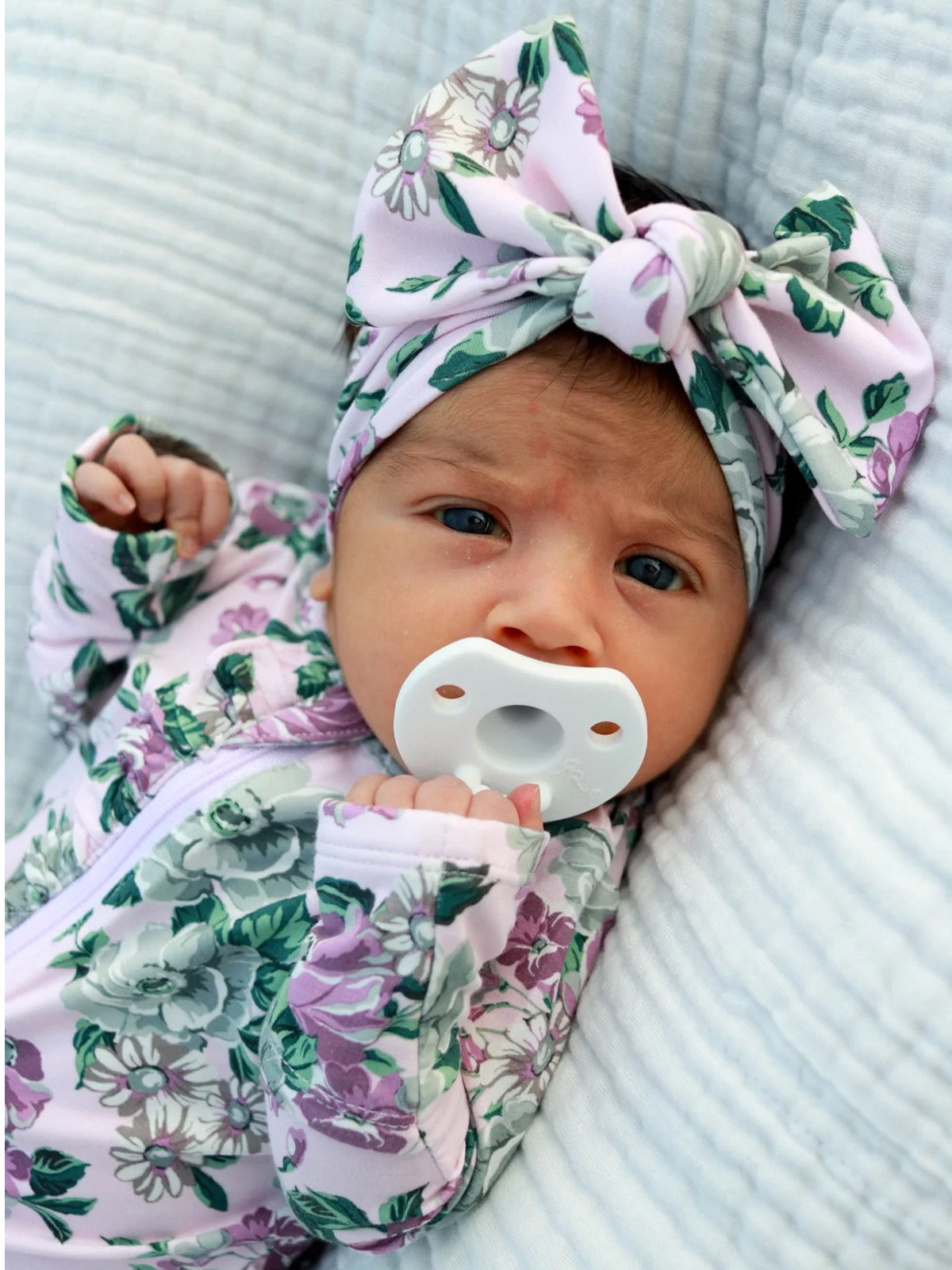 Infant with a floral onesie and a large bow headband, holding a pacifier, laying on a soft, textured surface.