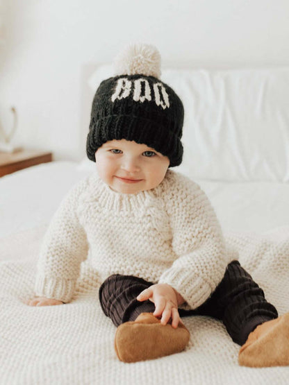 Smiling toddler in a cozy sweater and black beanie sitting on a white blanket.