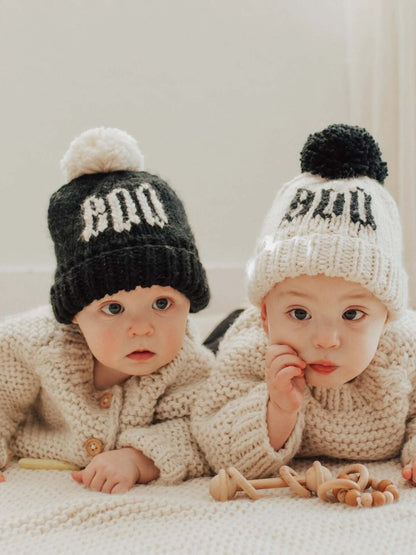 Two infants in cozy knit sweaters wear black and white pom-pom hats, engaged with wooden toys on a light background.