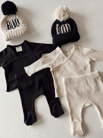 Two sets of baby clothing: black and cream ribbed tops and pants, paired with knit hats featuring "Boo" text.
