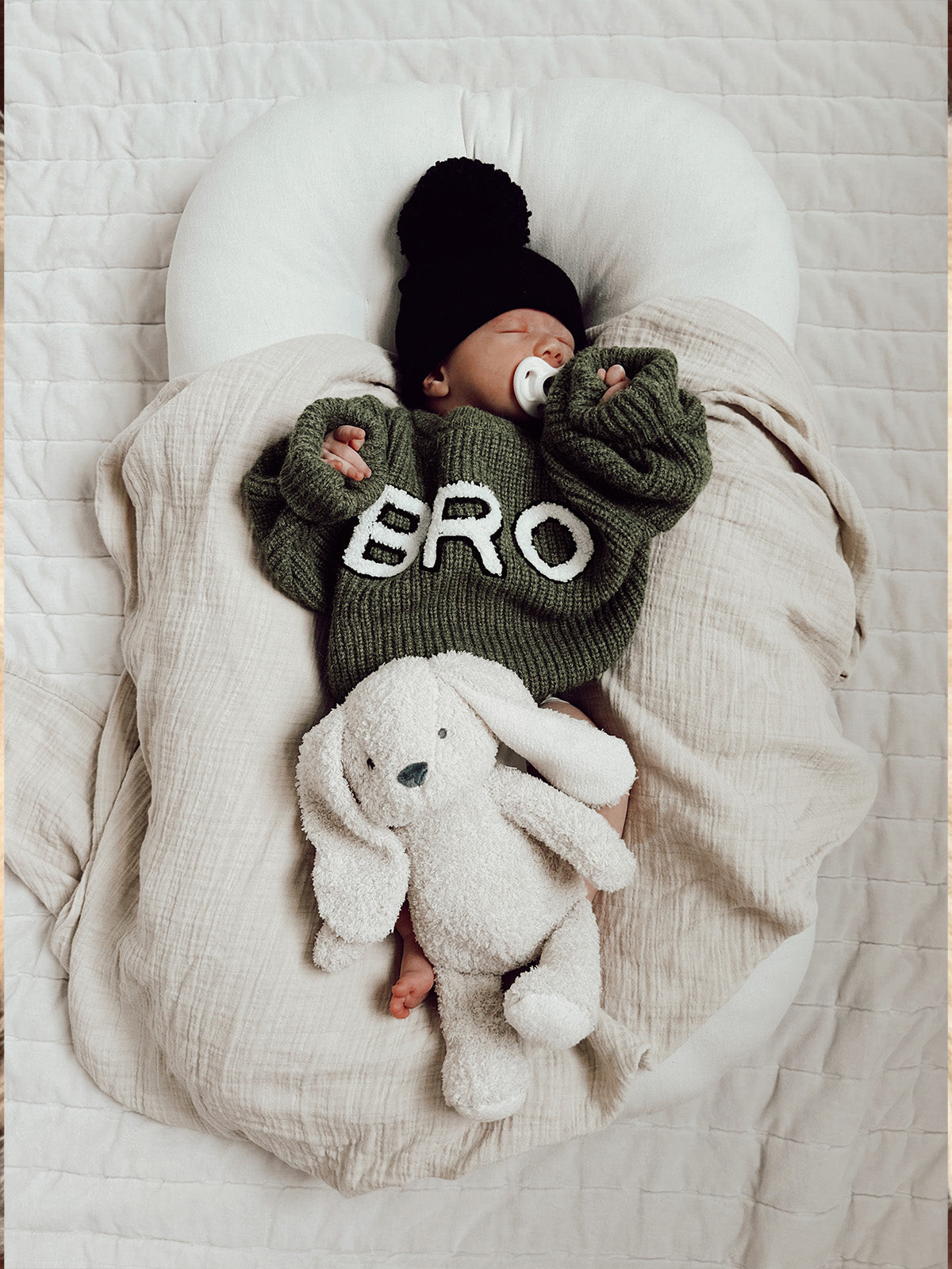 Infant in green sweater with "BRO" resting on a blanket, holding a white plush bunny. Cozy indoor setting.