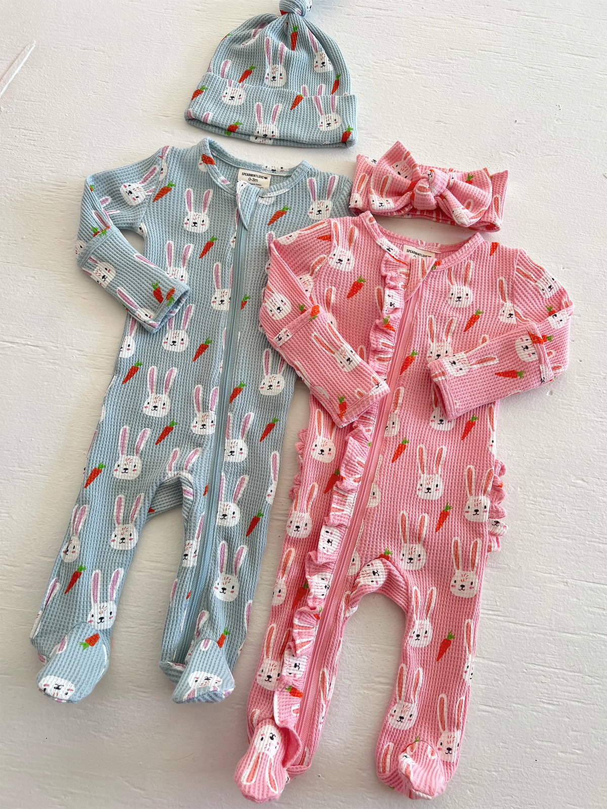 Two bunny-patterned baby onesies and matching hat and headband, in blue and pink, displayed on a white surface.