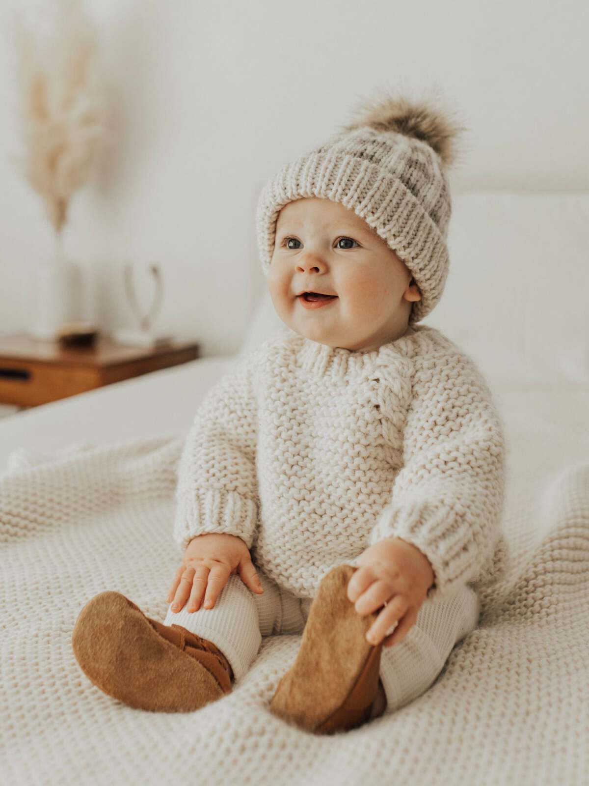 Smiling baby in a cozy knitted sweater and hat, seated on a soft blanket in a bright room.