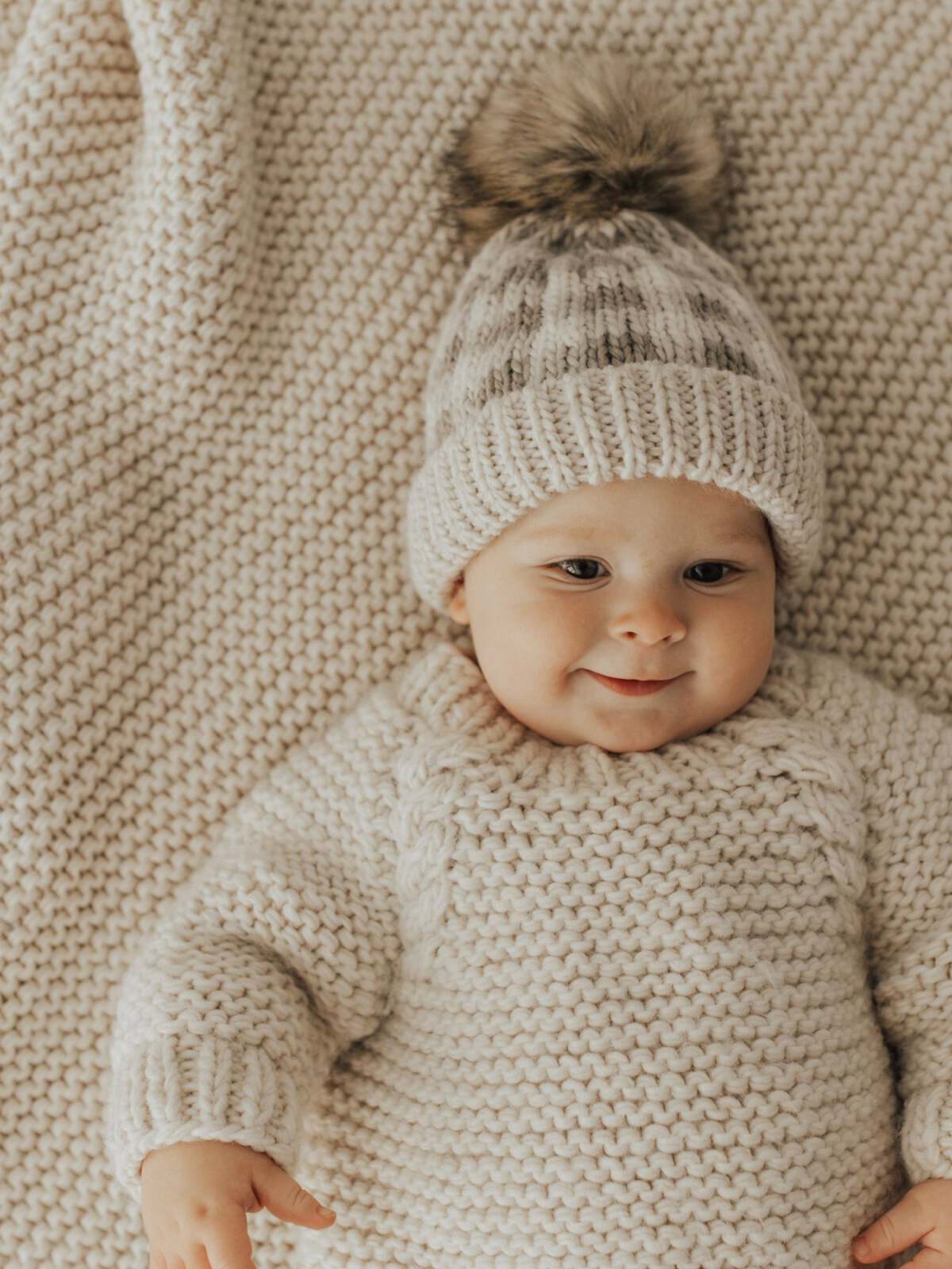 Smiling baby in a knitted sweater and hat on a textured blanket, creating a warm, cozy scene.
