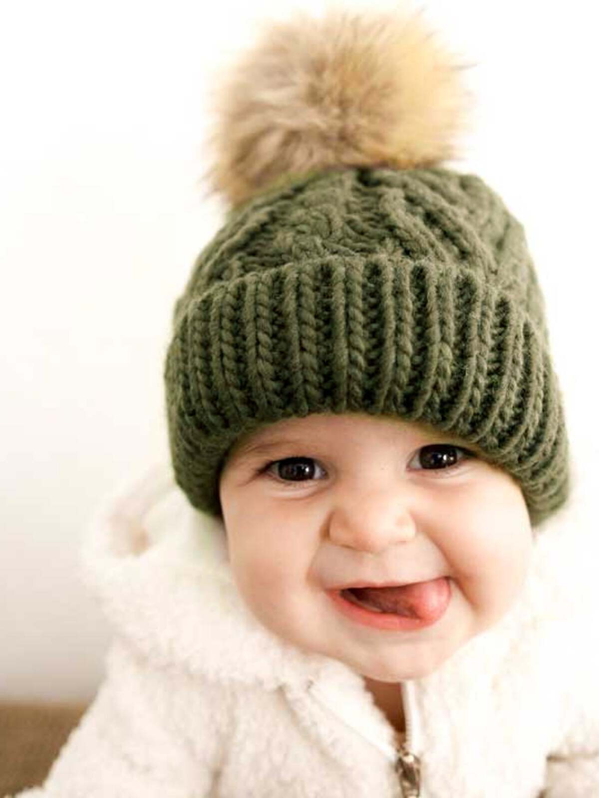 Smiling baby wearing an olive green knit hat with a pom-pom, sticking out tongue, and cozy white jacket.