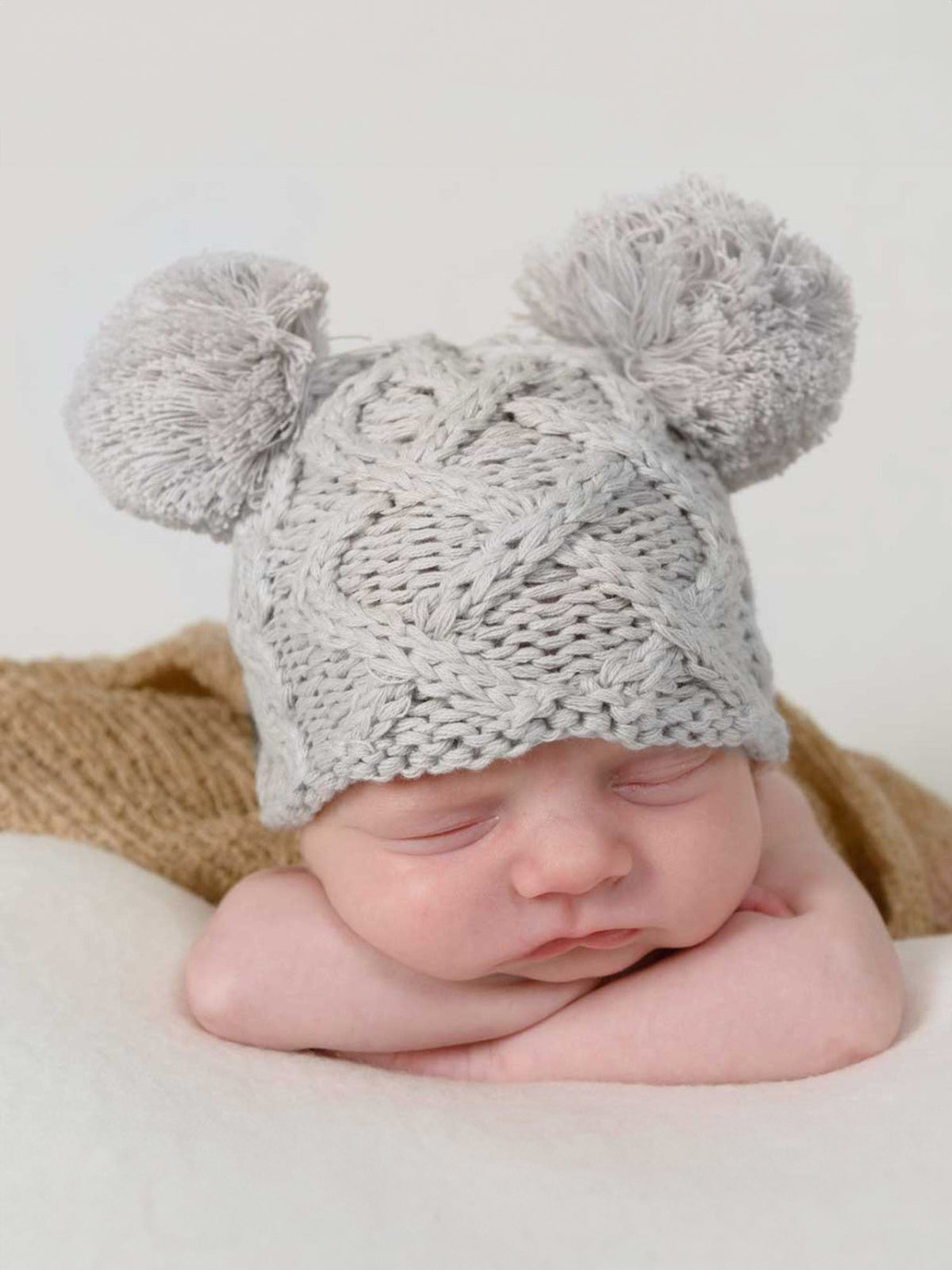 Newborn baby sleeping peacefully, wearing a cozy gray knitted hat with pom-poms. Soft textures and calm setting.