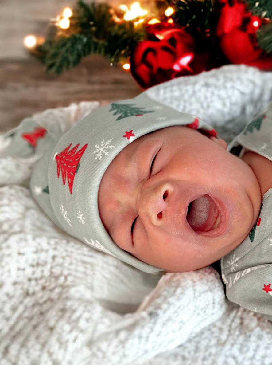 Newborn baby yawning in a cozy blanket, wearing a festive gray hat with Christmas tree patterns.
