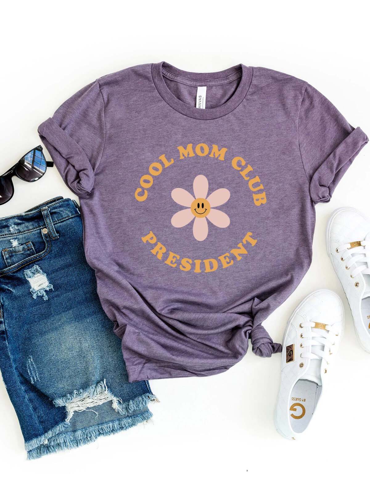 Purple t-shirt with "Cool Mom Club President" text, styled with denim shorts, sunglasses, and white sneakers.