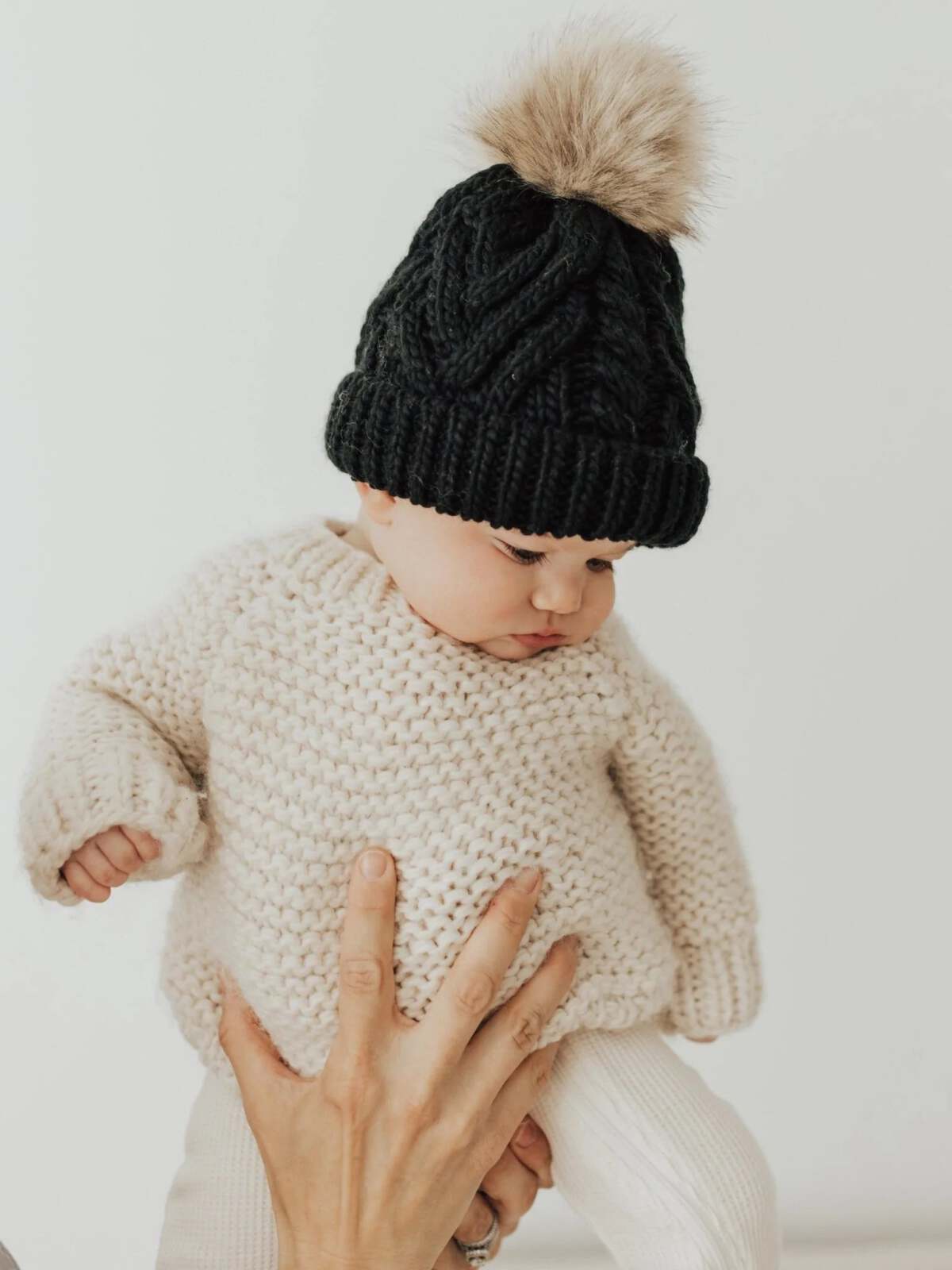 Baby in a cozy cream sweater and black knitted hat with a pom-pom, held by an adult hand.